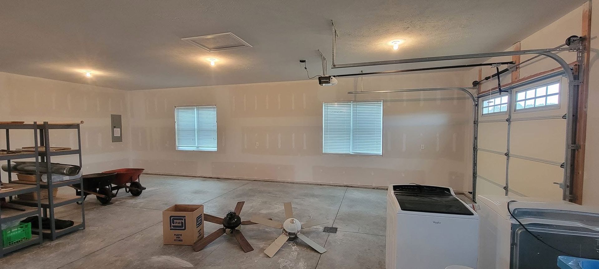An unfinished garage interior with drywall, windows, a partially open garage door, and scattered tools.