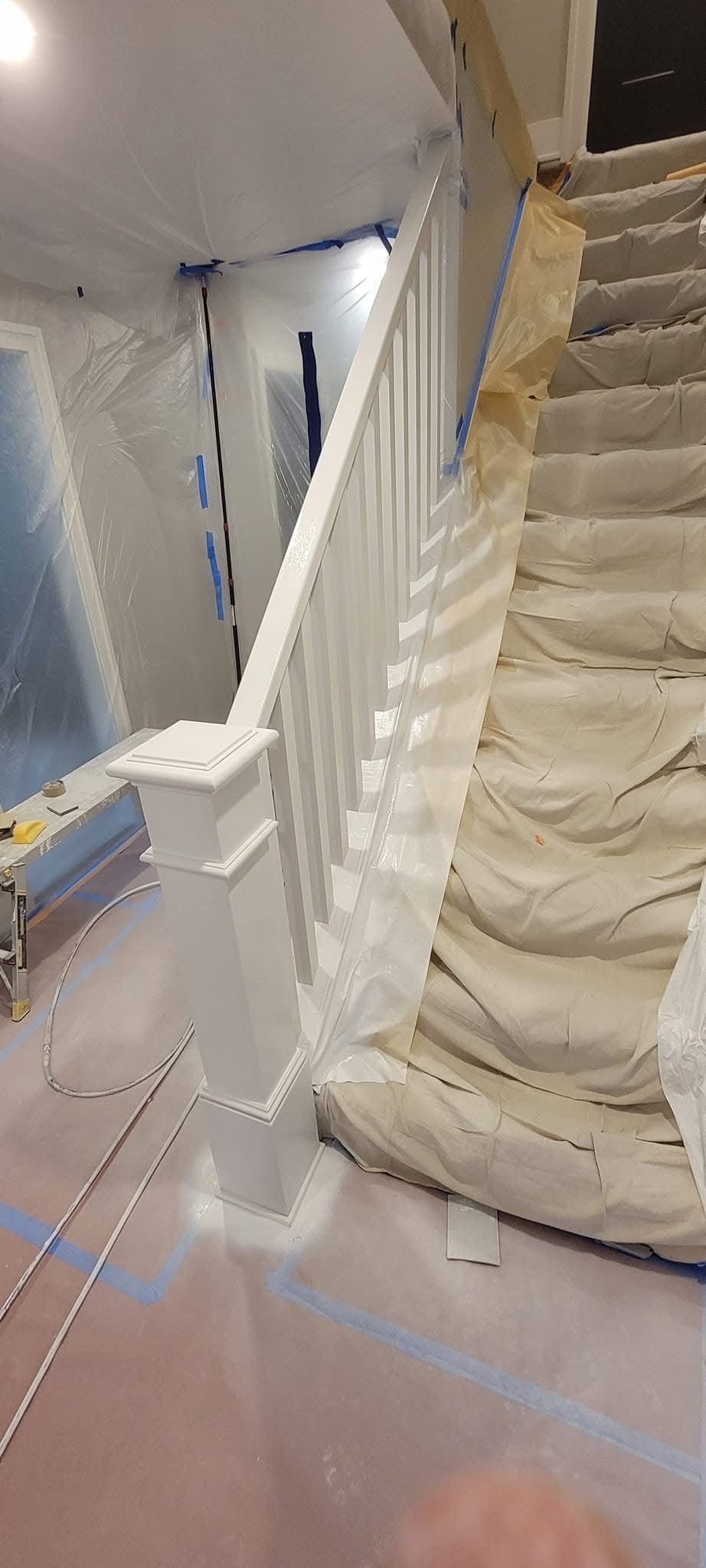A staircase being painted, covered in protective plastic and tape. White railing and stairs.