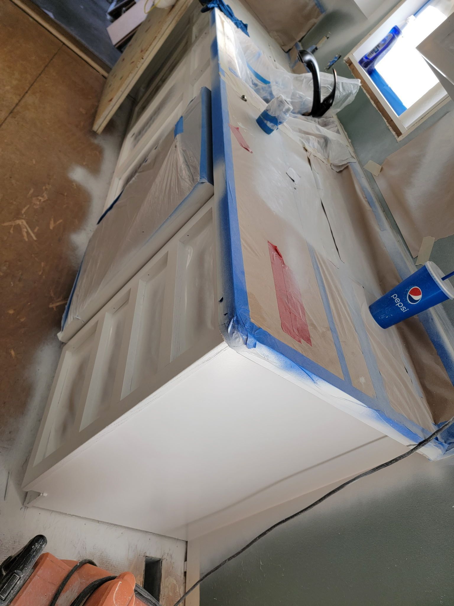 Kitchen cabinets covered in plastic and blue tape, being painted white.