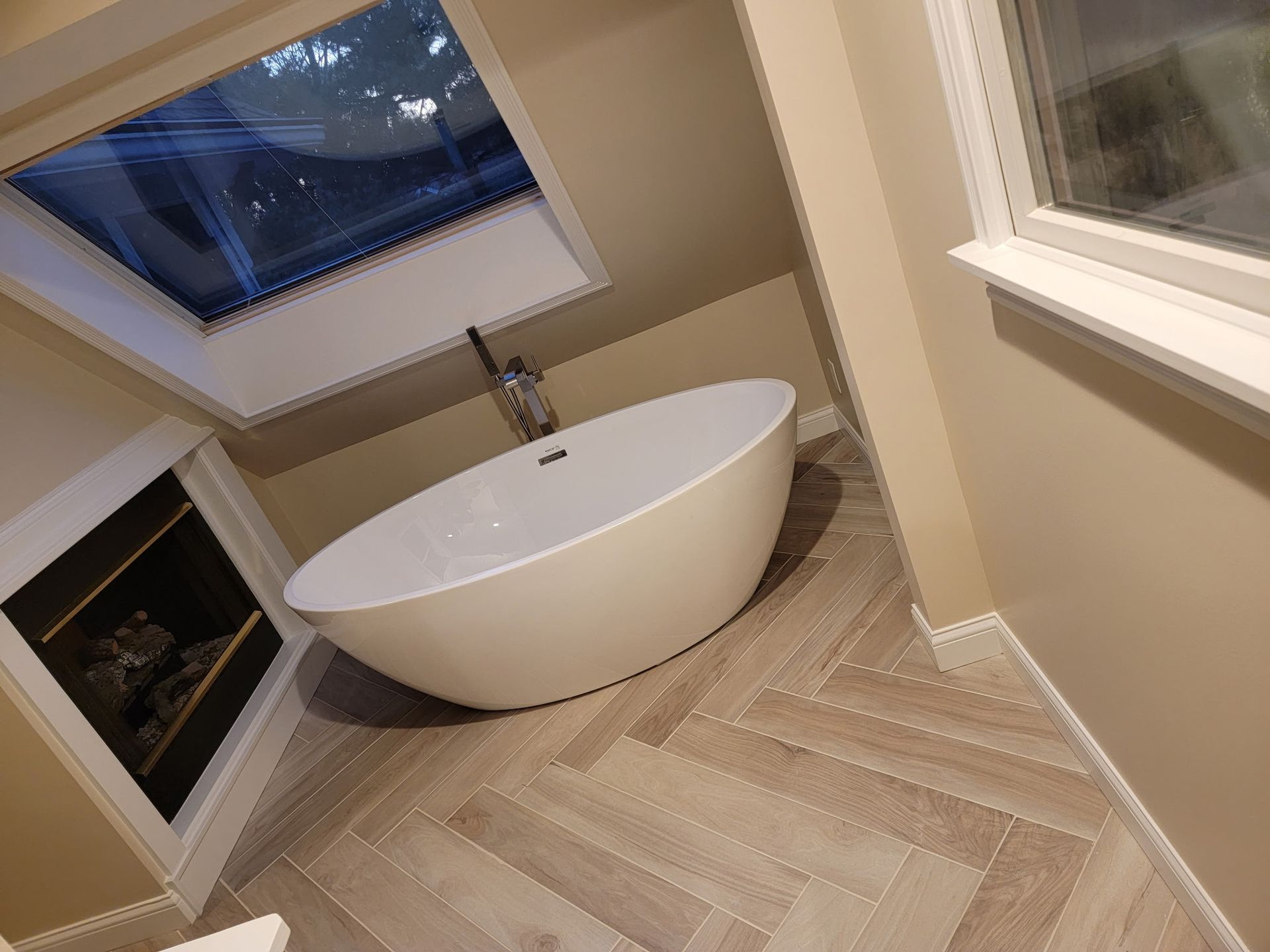 Bathroom with a white oval bathtub, skylight, and beige herringbone tile floor.