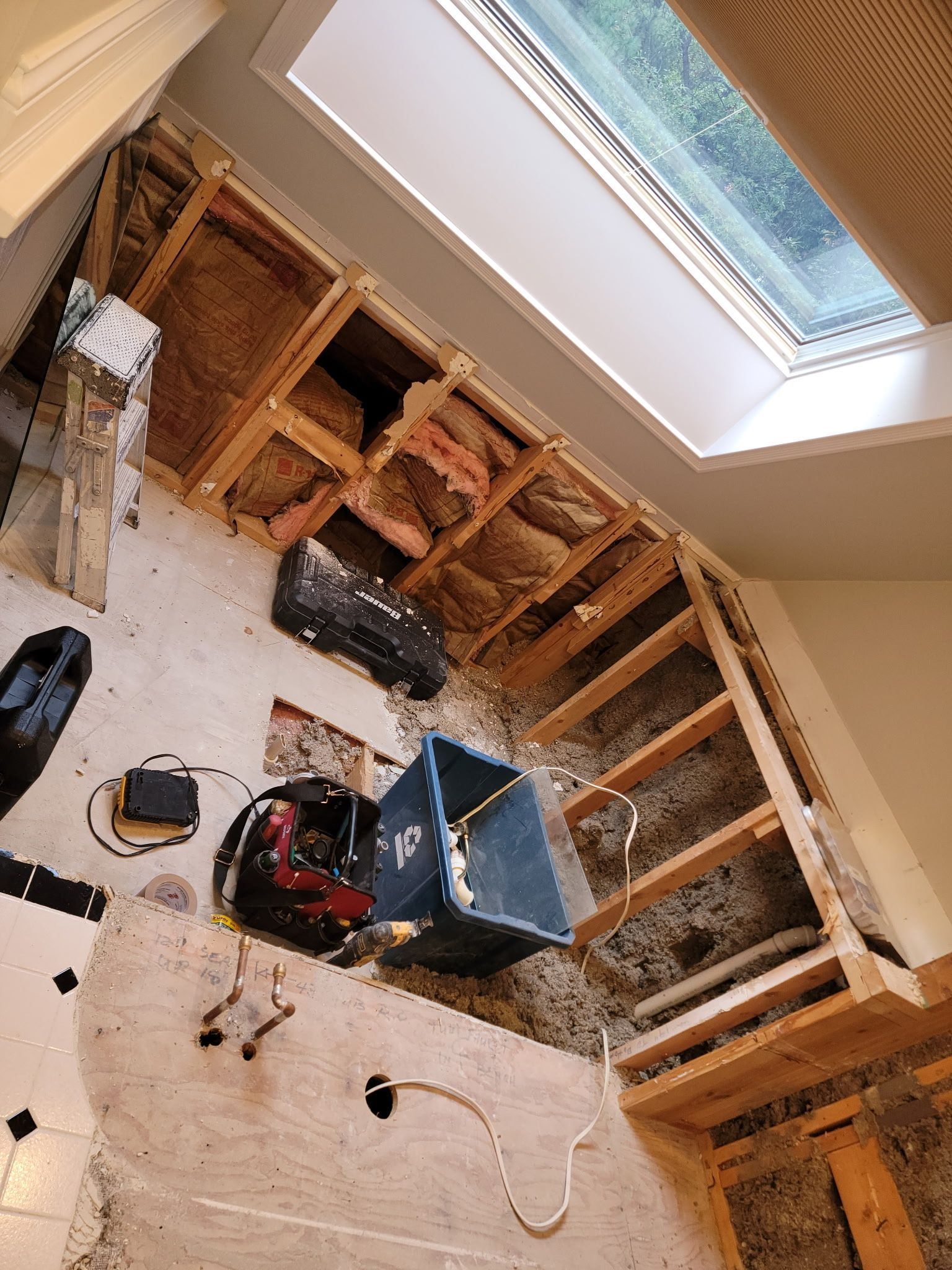 Interior wall under construction; exposed wood framing, insulation, and skylight. Blue bucket and tools on the floor.