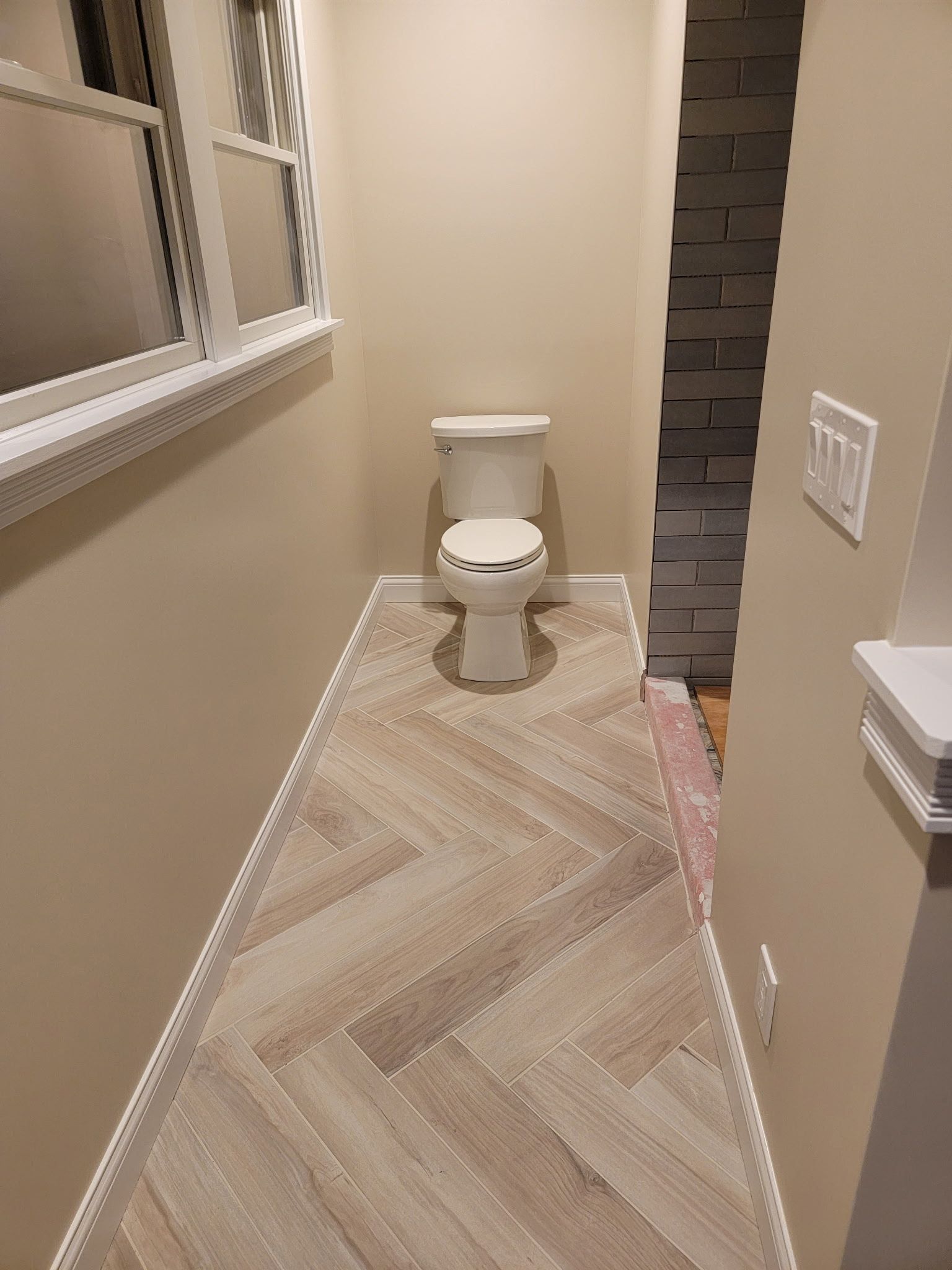 Narrow bathroom with herringbone tile floor, toilet, window, and stacked stone wall.