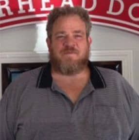 A man with a beard is standing in front of a sign.