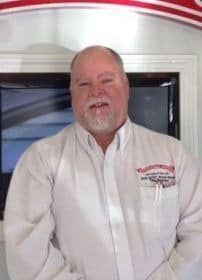 A man with a beard is wearing a white shirt and standing in front of a television.