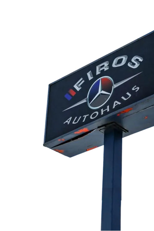 A dark, rectangular FIROS Autohaus sign mounted on a post against a white sky, featuring the Mercedes-Benz logo | Firos Auto House 2