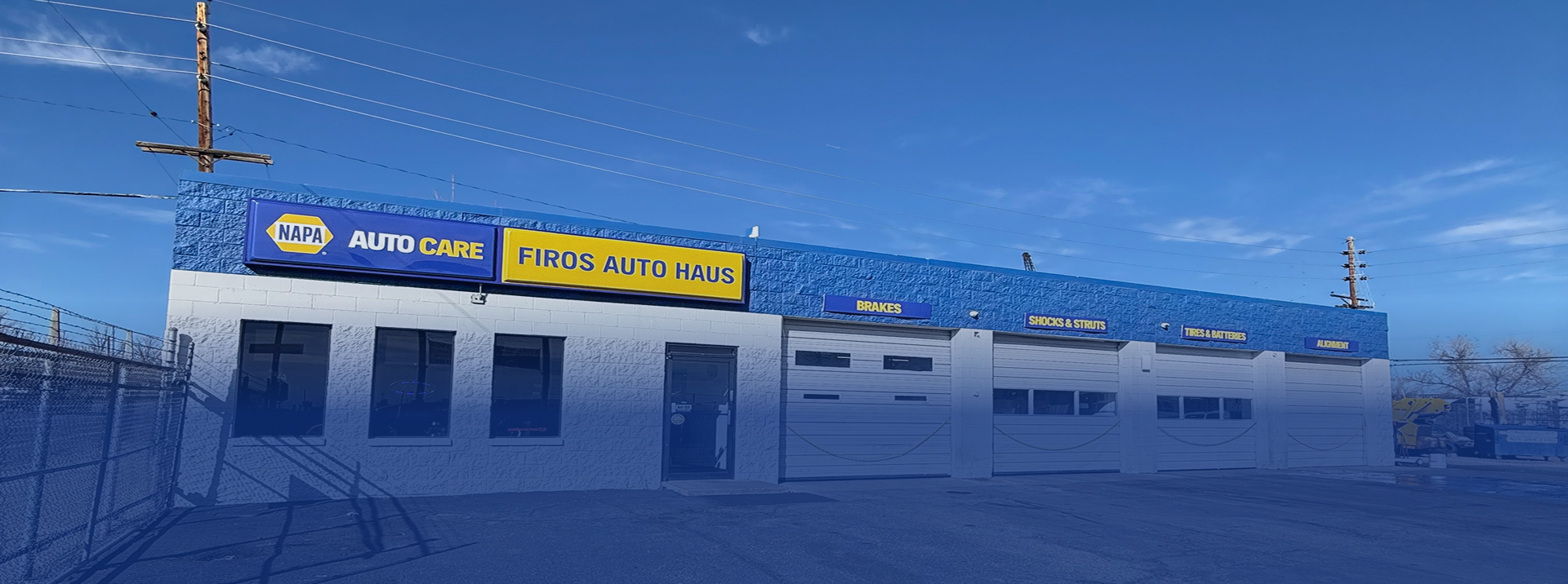 A NAPA Auto Parts store with a blue and white exterior, yellow signage, and several garage bays under a clear blue sky | Firos Auto House 2
