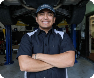 A smiling mechanic wearing a cap and uniform stands with arms crossed in a garage, with a car lifted on a rack above | Firos Auto House 2