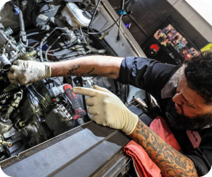 A tattooed mechanic in gloves works on an open car engine, pointing toward a specific component with one hand | Firos Auto House 2