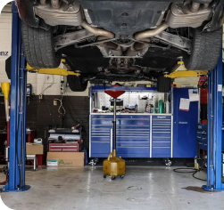 A car elevated on a two-post lift in a garage, with a mobile oil drain cart positioned underneath | Firos Auto House 2