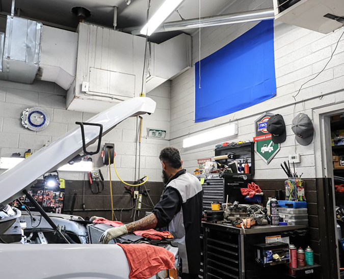 A mechanic works on the engine of a white vehicle inside a garage with tools, shelves, and a blue flag on the wall | Firos Auto House 2