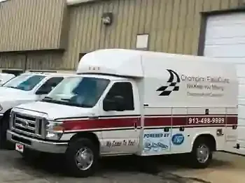 A worker in a plaid shirt and hat inspects the tires of a large blue semi-truck parked in a lot.