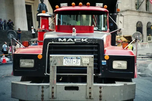 Front view of a red and black Mack dump truck parked on a city street, featuring a large metal bumper and license plate.