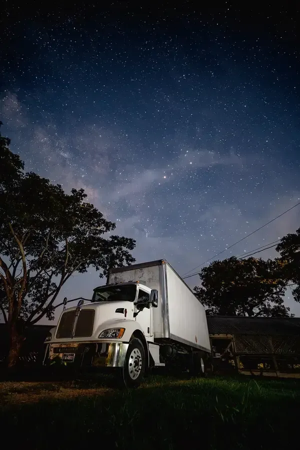 A white box truck parked under a vibrant, starry night sky with silhouettes of trees on the sides.