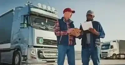 Two people in vests and hats stand in a truck yard, reviewing documents in front of a parked semi-truck.