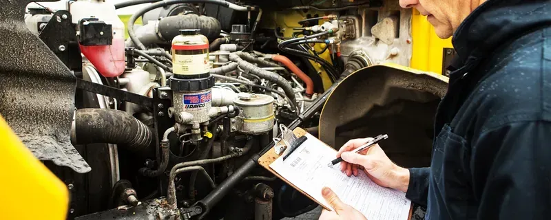 A mechanic in a dark uniform writing on a clipboard while inspecting an open engine compartment of a yellow vehicle.