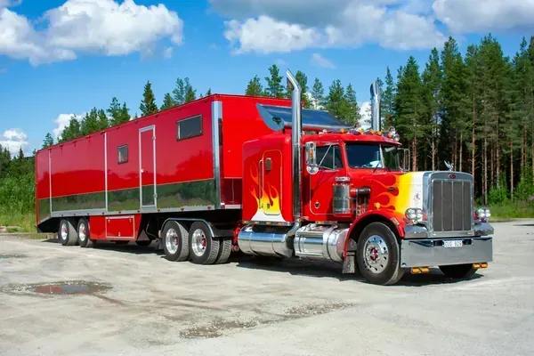 A bright red semi-truck with flame decals on the hood, hauling a long red trailer, parked on a dirt lot near a forest.