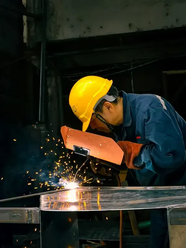 A worker in a yellow hard hat and blue uniform uses a handheld welding shield while sparks fly from a metal workspace.