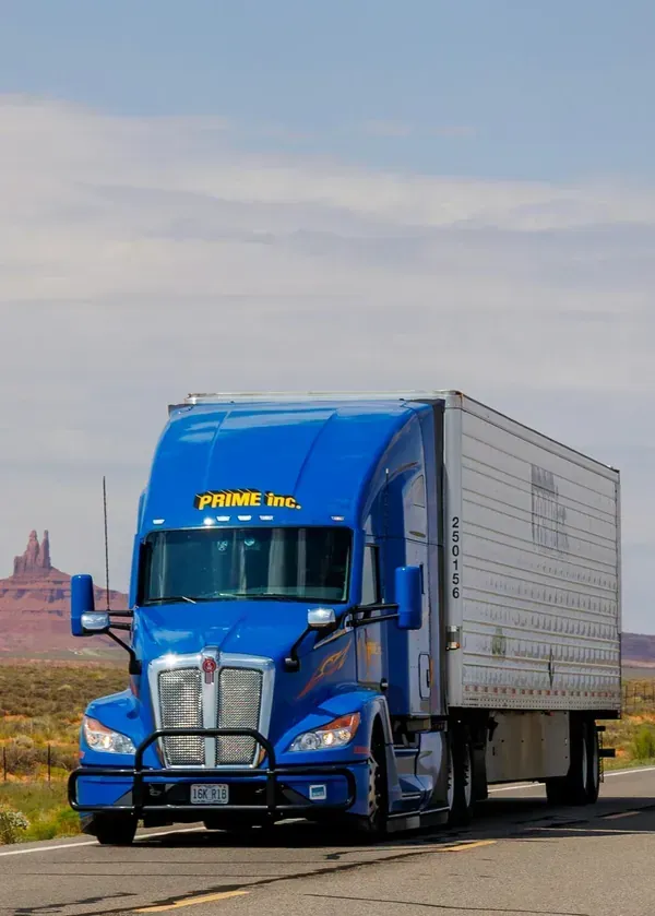 A blue Prime Inc. semi-truck driving on a road in the desert near rock formations.