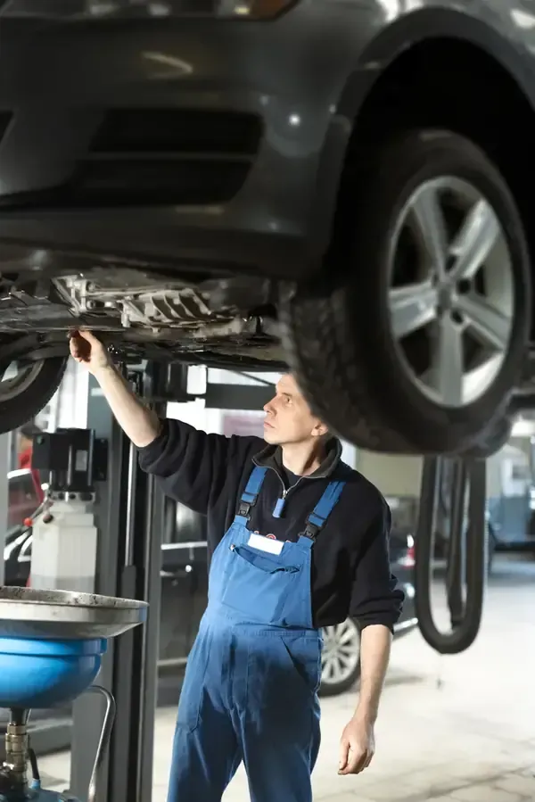 A technician in blue overalls works under a car raised on a hydraulic lift in an automotive workshop.