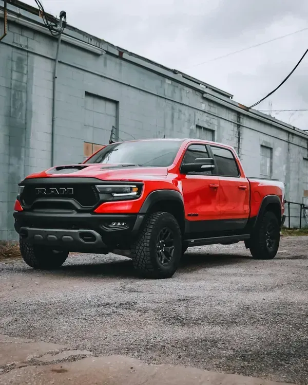 A bright red Ram TRX pickup truck parked on a gravel lot in front of a weathered, grey industrial building.