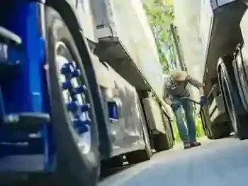 A worker in a plaid shirt and hat inspects the tires of a large blue semi-truck parked in a lot.