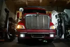 A red semi-truck parked in a garage, illuminated by its yellow headlights and white bumper fog lights.