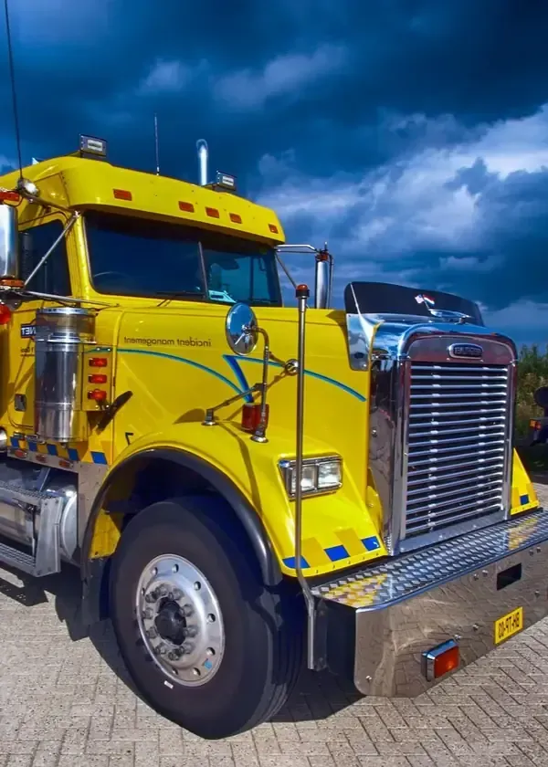 A bright yellow Freightliner semi-truck parked on a cobblestone surface under a dramatic, dark, cloudy sky.