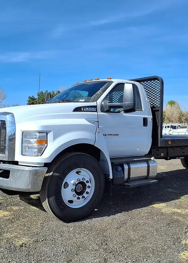 A white Ford F-650 flatbed truck parked on a gravel lot under a clear blue sky.