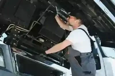 A person wearing a white t-shirt and grey overalls works on the engine of a large vehicle.