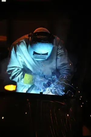 A welder wearing protective gear and a mask works on metal, producing a bright blue electric arc and glowing sparks.