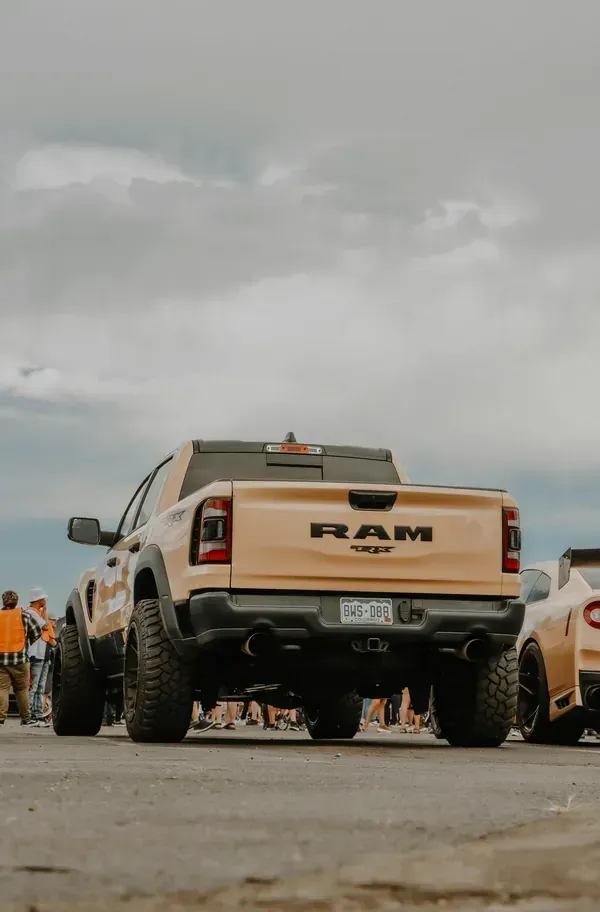 A tan RAM truck parked outdoors, with a partial view of another vehicle and a blurry crowd in the background.