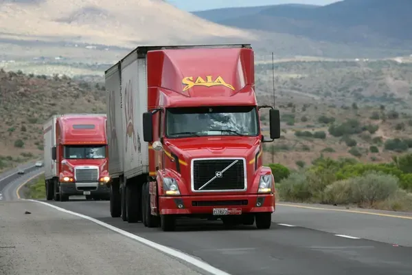 Two red semi-trucks with white trailers drive along an open road in a dry, mountainous landscape.
