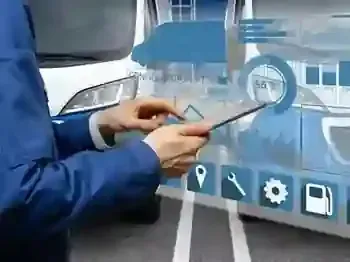 A worker in a plaid shirt and hat inspects the tires of a large blue semi-truck parked in a lot.