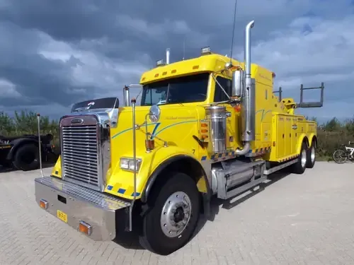 A bright yellow Freightliner tow truck parked on a paved lot under a cloudy sky.