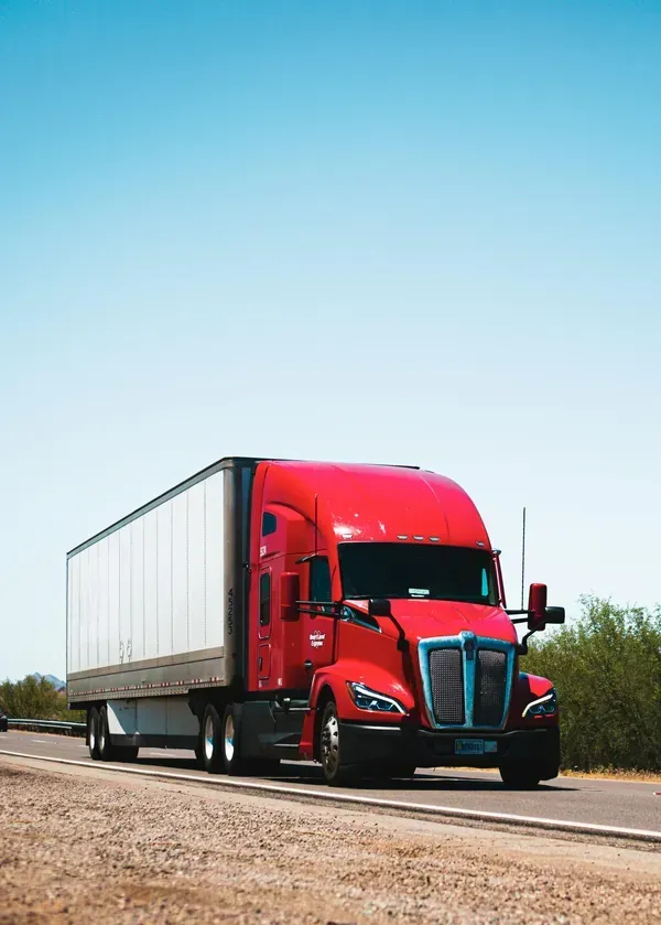 A red semi-truck with a white trailer drives along a rural road under a clear blue sky.