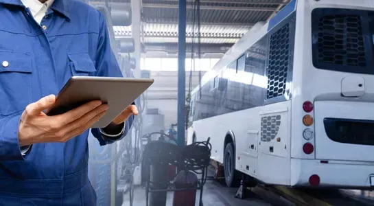 A person in a blue uniform holds a tablet in front of a white bus inside a service garage.