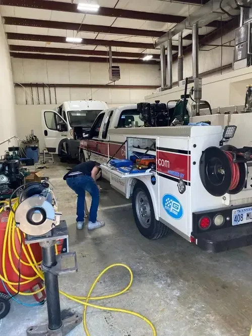 A person works on a white service truck inside a garage, with another vehicle parked in the background.