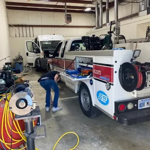 A person works in a garage on a white utility truck with red stripes, parked near a van and shop equipment.