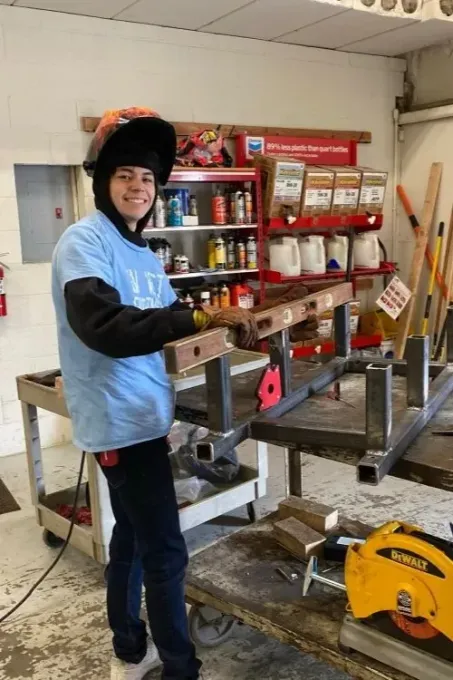 A smiling person in a blue shirt and welding hood holds a level on a metal frame inside a workshop.