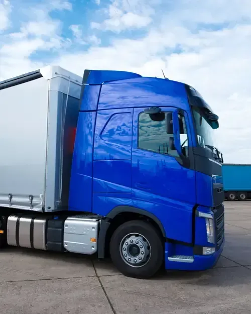 A side profile of a bright blue semi-truck cab with a silver trailer parked on a concrete lot under a cloudy sky.
