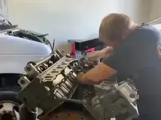 A person works on a large automotive engine suspended from an engine hoist in a garage.