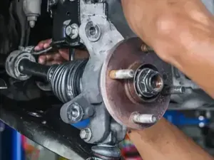 A mechanic's hands work on the front suspension and wheel hub assembly of a car in a repair shop.
