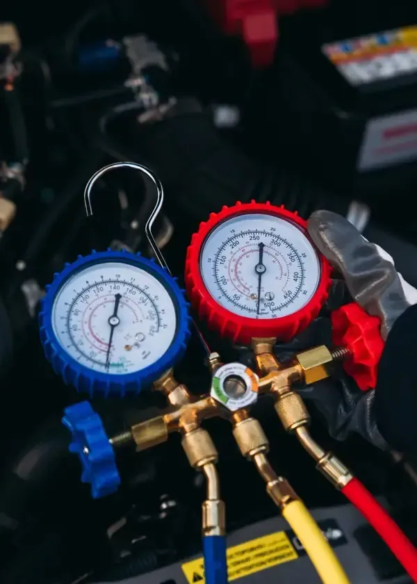 A technician’s gloved hand holding a manifold gauge set with red and blue dials to check a vehicle’s air conditioning.