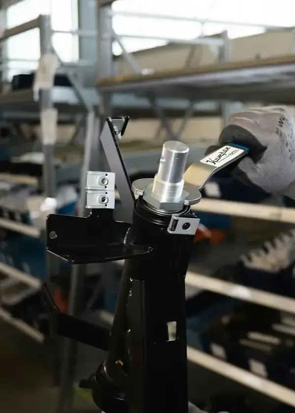 A gloved hand uses a specialized metal tool to tighten a silver fitting on a black industrial component in a warehouse.