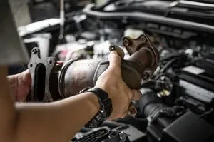 A mechanic holding a catalytic converter in an engine bay.