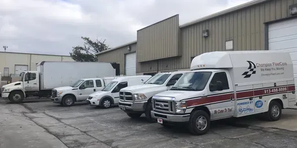 A line of white service vehicles parked in front of a tan industrial warehouse building under a cloudy sky.