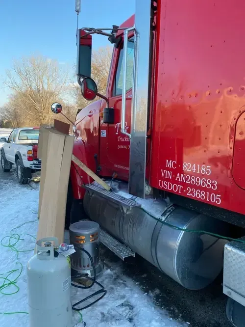 A red semi-truck parked in snow, with a portable propane heater and cardboard structure set up near the driver's door.