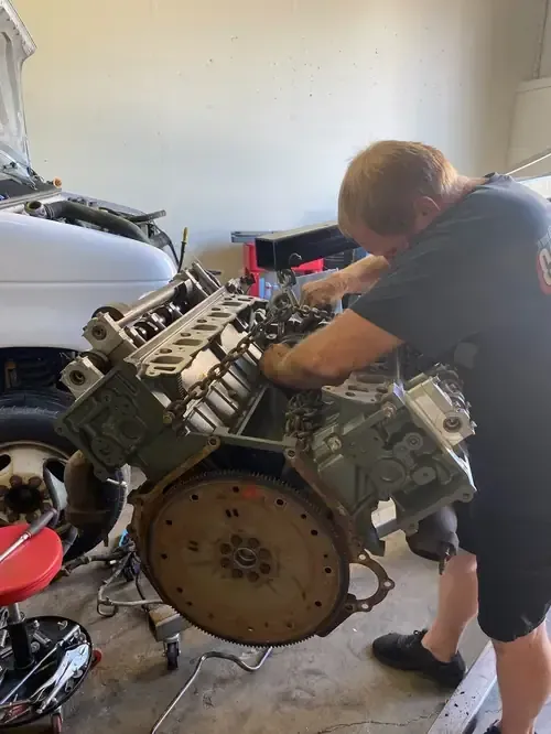 A person works on a large automotive engine suspended from an engine hoist in a garage.