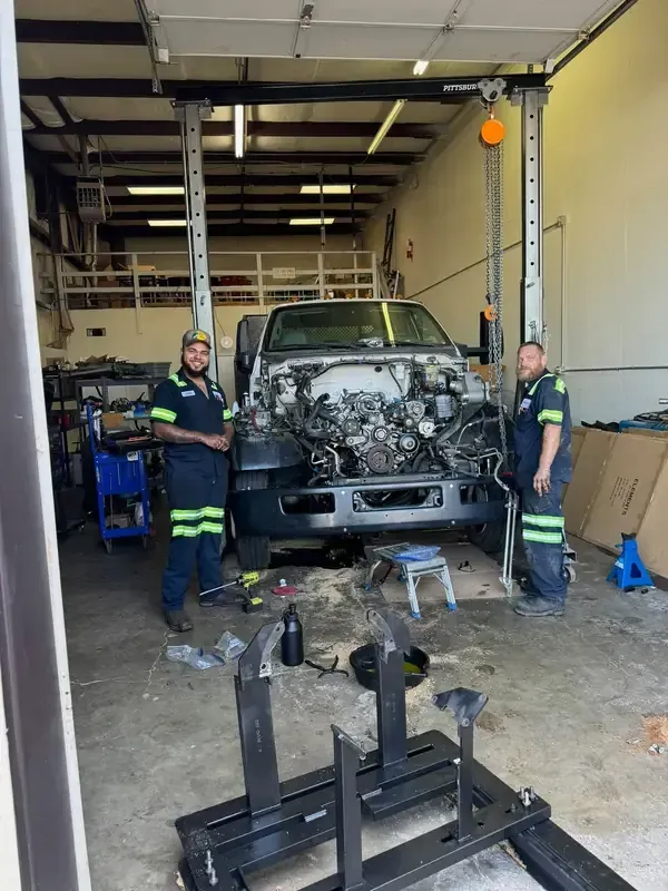 Two mechanics stand in an auto repair shop with a truck hoisted up, its engine compartment exposed for service.