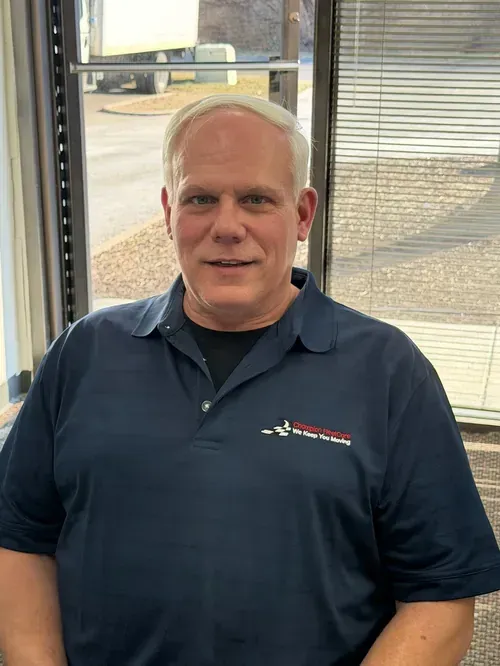 A person wearing a dark blue polo shirt with a company logo, standing indoors in front of a window with blinds.